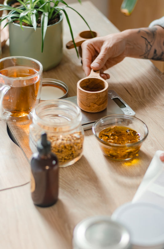 A person's hands holding a small bowl of herbs with a glass of tea and a bottle in the foreground on a wooden surface, likely related to herbal wellness咨询.